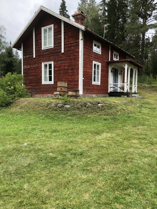 Rött trädhus med vita knutar och förstukvist, omliggande grön gräsmatta och skog. Markförändring syns vid nedre hörnet. Gamla tunnor är placerade nära huset.