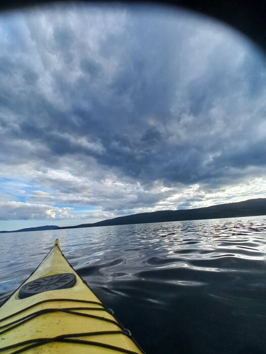 En bild av spetsen på en gul kajak på en lugn sjö med mörka moln på himlen och skogsklädda kullar i bakgrunden.