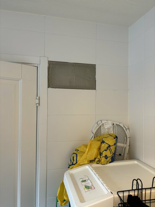 Vägg i tvättstuga där kakelplattor har släppt, vilket exponerar fogen bakom. Nedanför syns en tvättmaskin med en gul plastpåse och en ihopvikbar torkställning.