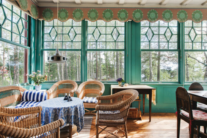 Inglasad veranda med rottingmöbler och blåvita textilier, bord med blå duk, utsikt mot skog och sjö, gröna fönsterkarmar och mönstrad gardinvalans.