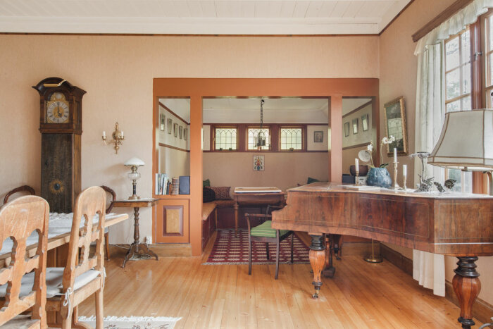 Ett vardagsrum med trägolv, en stor gammal klocka, ett matbord med stolar, en flygel och en mysig sittgrupp med kuddar i en alkoverk.