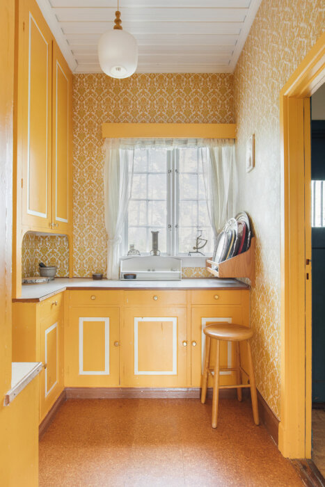 Gult retro-kök med vita detaljer, diskbänk och fönster med vita gardiner. Köket har en pall och flera brickor uppställda på diskbänken.