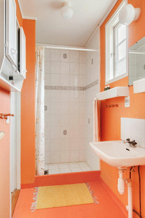 Badrum med vit kaklad dusch, orange målade väggar och golv, samt vitt handfat bredvid ett fönster med dagsljus.