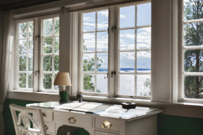Utsikt från ett rum med ett skrivbord framför stora spröjsade fönster, som vetter mot en glittrande sjö och ett bergigt landskap i fjärran.