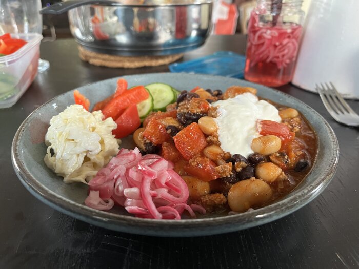En tallrik med chili con carne toppad med gräddfil, picklad rödlök, coleslaw, gurka och paprika samt tillbehör i bakgrunden.