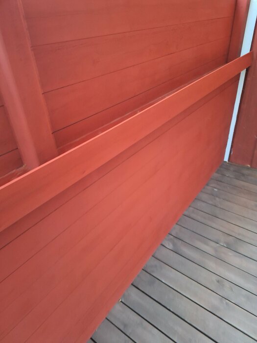Nymålad yttervägg i en stark röd färg, del av husets fasad och en beskuren vy av verandagolv i brunt trä.