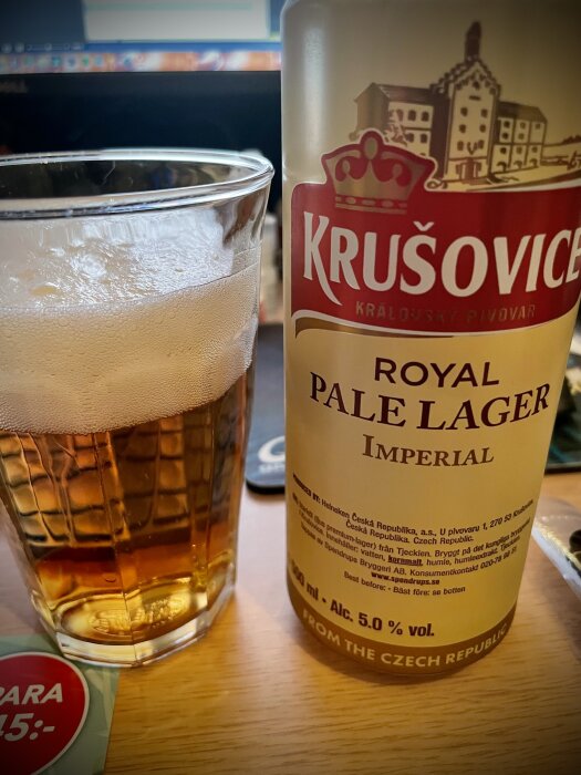 En glas öl bredvid en flaska Krušovice Royal Pale Lager Imperial med 5,0% alkoholhalt på ett skrivbord.