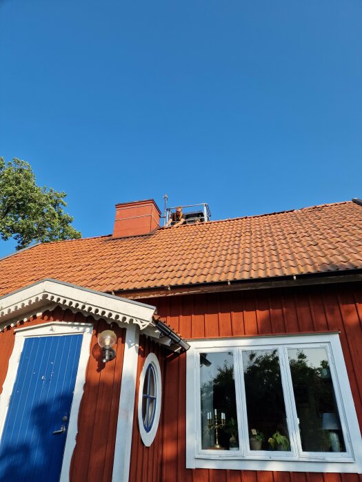 Röd träfasad med vita detaljer på ett hus med tegeltak och blå ytterdörr. En person står på taket nära skorstenen, under en klarblå himmel.