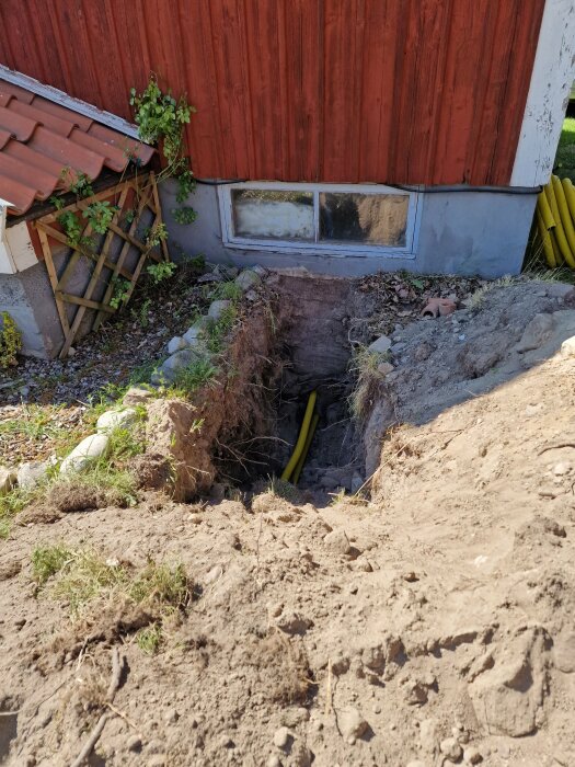 Grävt dike vid sidan av ett hus med röd träpanel, med gul kabel synlig i diket och ett fönster till en källare i bakgrunden. Växter och byggmaterial till vänster.