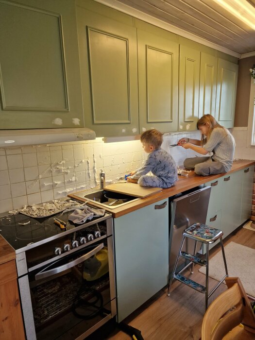 Två barn tar bort kakel i ett grönt kök, sittande på köksbänken. Köket är under renovering och verktyg ligger utspridda. En stege står bredvid diskmaskinen.