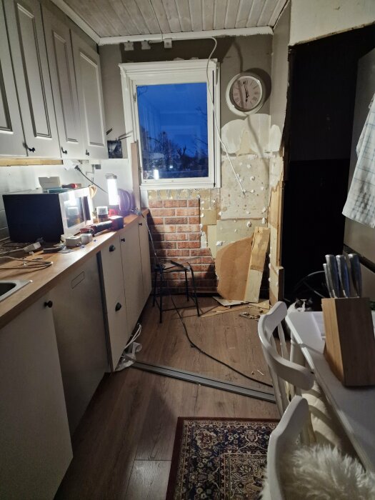 Ett kök under renovering med verktyg och byggmaterial på bänken och golvet. Väggpaneler saknas och en lampa lyser på ett köksskåp.