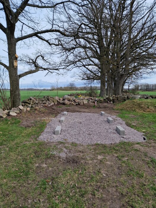 Grund för ny byggnad med fyra betongblock på grus och underligtgande jord, omgiven av träd och stengärdsgårdar på landsbygden.