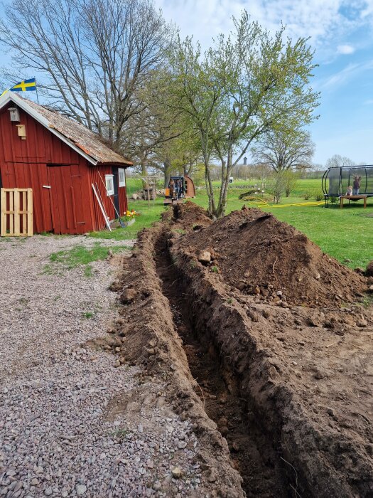 En grävmaskin som gräver ett dike vid sidan av ett rött hus med en svensk flagga. I bakgrunden finns träd, en gräsmatta och en studsmatta.