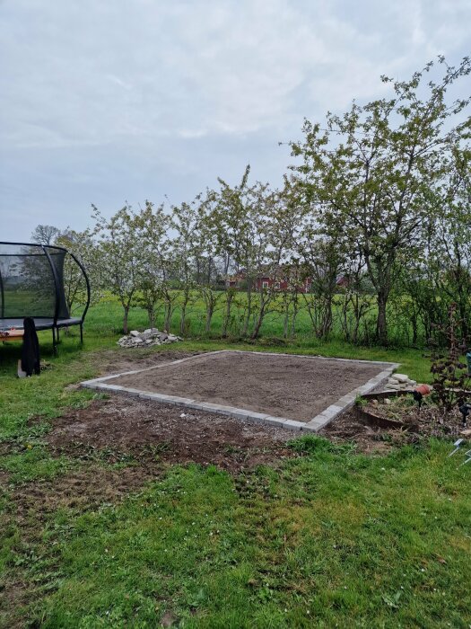 Kvadratisk yta förberedd för projekt i trädgård, omgiven av häckar med ett rött hus i bakgrunden. Trampolin synlig till vänster.