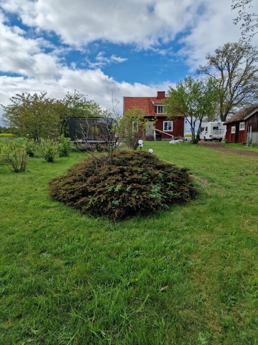 En lantlig trädgård med grön gräsmatta, buske i förgrunden, rött trähus med vita knutar i bakgrunden. Hoppa, båttrailer och husvagn syns också.