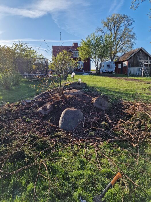 Jordhög omringad av stenar och grenar i en trädgård med ett rött hus och en husvagn i bakgrunden. Lummigt med gröna träd och blå himmel.