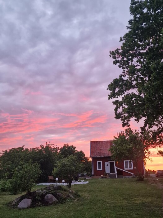 Rött hus vid solnedgång, omgiven av träd och en klippt gräsmatta. Barrikader och belysning syns i förgrunden, med en dramatisk rosa och lila himmel ovanför.