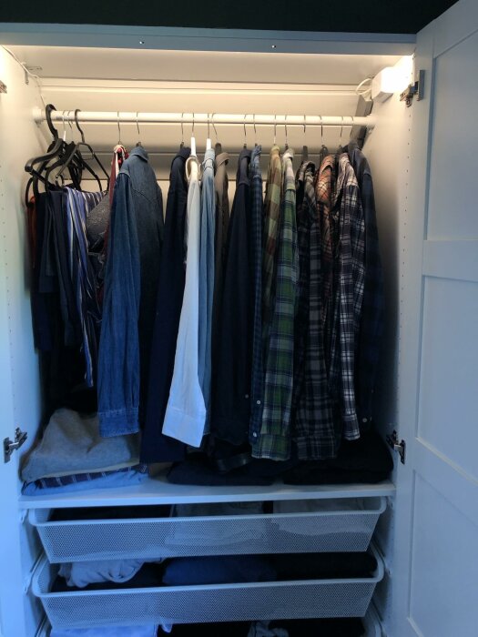 Garderoob med kläder på galgar och tre utdragbara trådkorgar, med inbyggd belysning upptill som lyser upp garderobsinnehållet.