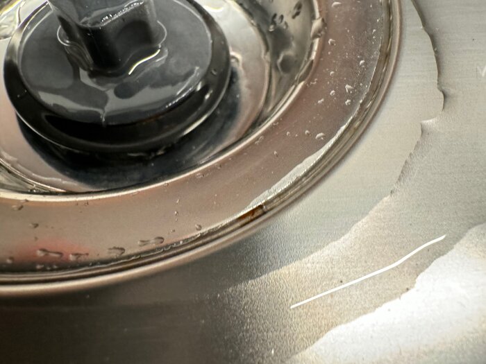 Närbild på en diskho i rostfritt stål med synlig rost runt bottenventilen och silen, samt en svart plastplugg.