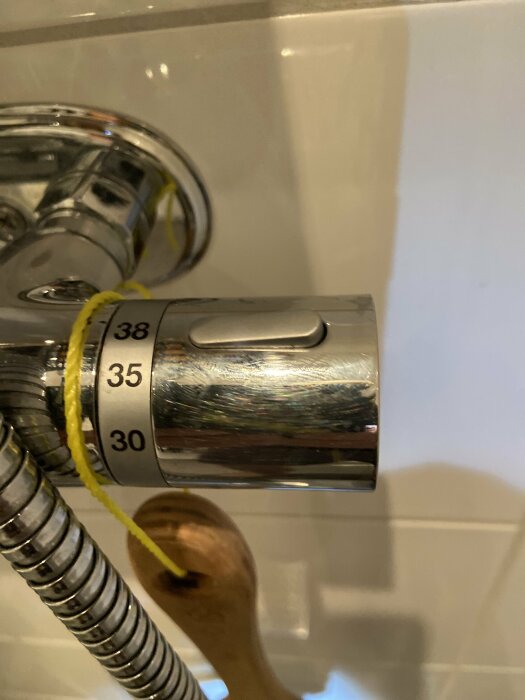 Duschtermostatinställning med temperaturskala från 30 till 38 grader och en knapp eller spärr för temperaturkontroll. Gula snören och duschslang synliga.