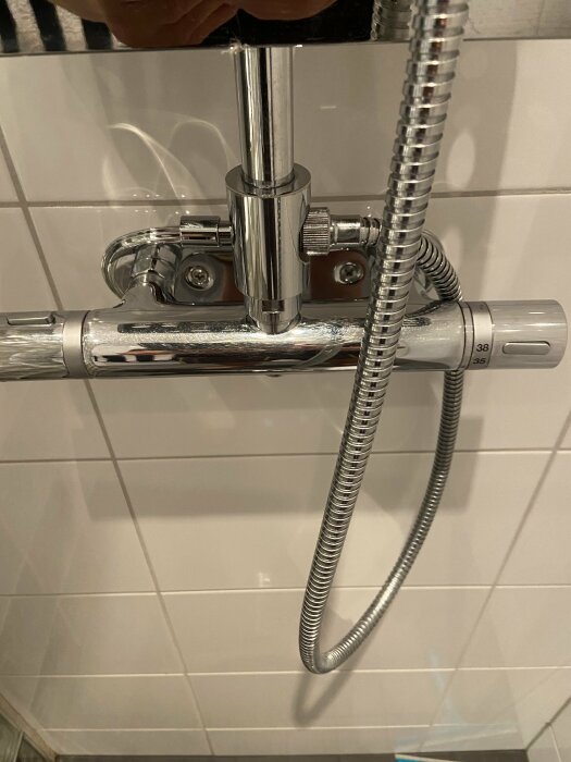 Närbild på en duschtermostat med en slang samt vred för att justera vattentemperaturen, monterad på vitkaklad badrumsvägg.