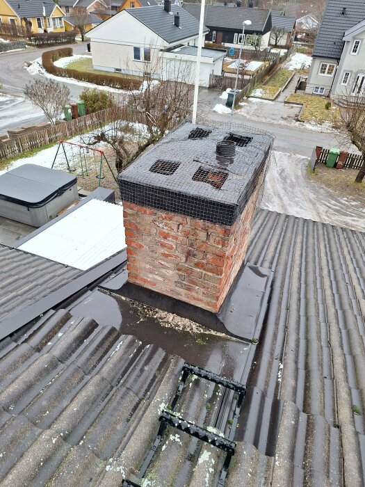 Sliten röd tegelsten-skorsten på ett hustak, utan fogar, täckt med ett nät. Omkringliggande områden är snötäckta, bilden är från en villakvarter.