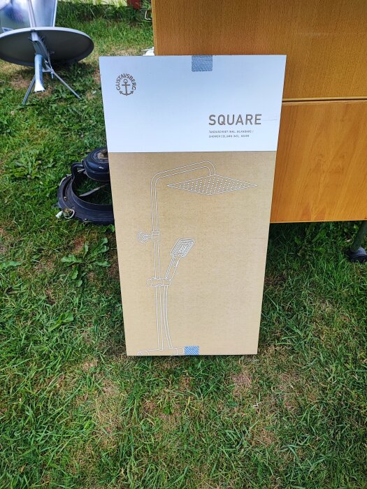 Förpackning av duschset från Gustavsberg modell Square, ståendes på gräsmatta framför en träskiva, redo för installation.