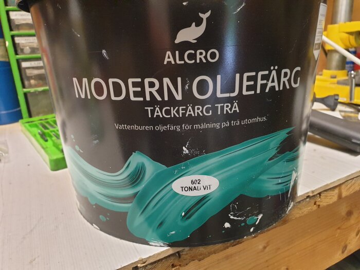 En burk Alcro Modern Oljefärg Täckfärg Trä 602 Tonad Vit för utomhusbruk står på en arbetsbänk i en verkstadsmiljö.