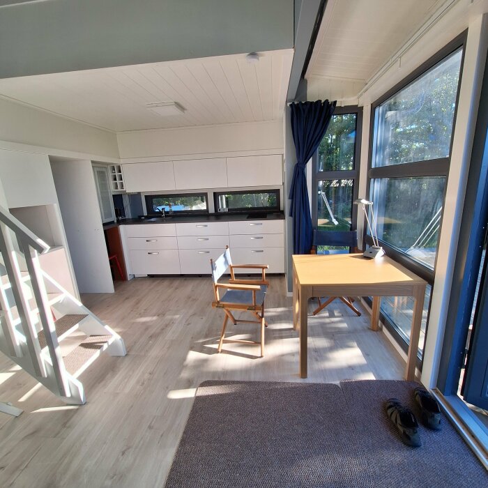 Nyrenoverad stuga med vitt kök, trappa, bord med stol och fönster med blå gardiner. Bild tagen innan möblering, ljusinsläpp från stora fönster.