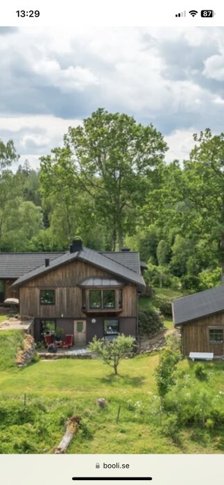 Trähus med svarta tak, äppleträd i förgrunden och elstängsel synligt längst ner, omgiven av grönska under en molnig himmel.