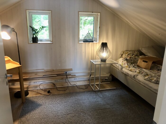 Loft med vit träpanel, två fönster, säng med lampa på vitt bord, skrivbord med skrivbordslampa och planta på fönsterbräde. Byggmaterial på golvet.