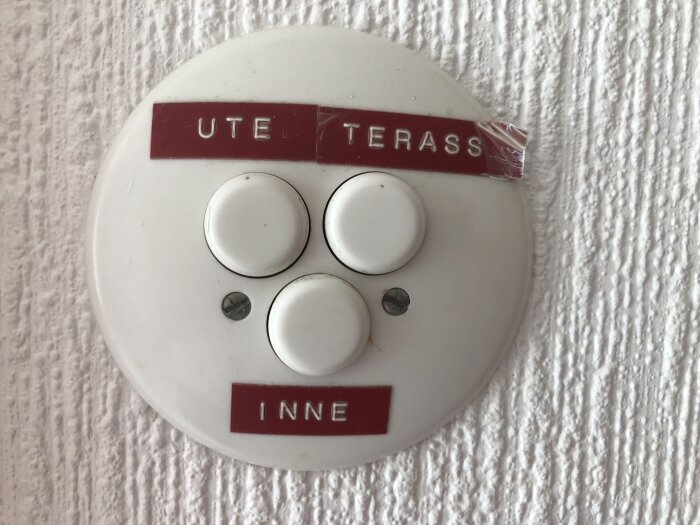 En rund vit knappanel med märkena "UTE", "INNE" och "TERASS" på en vit strukturerad vägg.