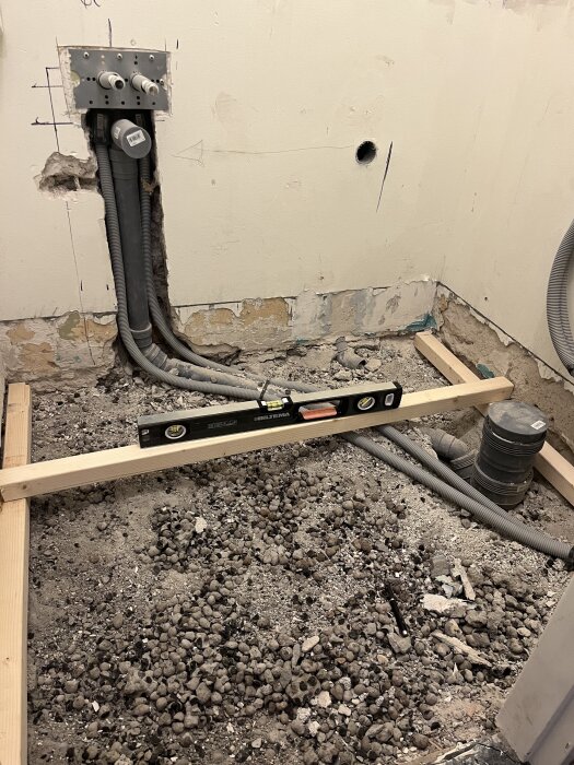 Renovering av badrum med öppnat golv, synliga vattenledningar och avloppsrör. En träregel med vattenpass visar den tilltänkta höjden på betongplattan.