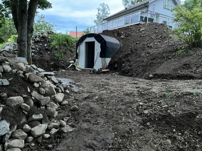 En jordkällare med välvt tak och stöttad framsida under byggnation, omgiven av jordhögar och stenar, med ett hus i bakgrunden.