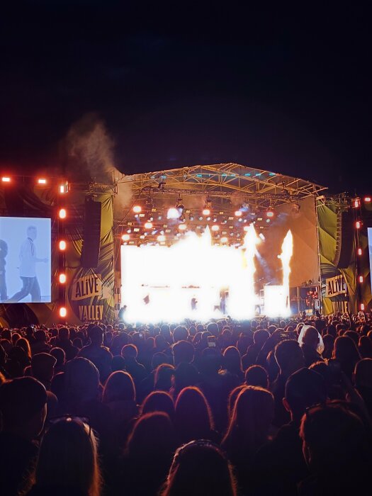 Stor publiksamling framför en scen på en musikfestival på natten, med pyroeffekter och ljusshow på scenen. En bildskärm till vänster visar en projektion av artisten.