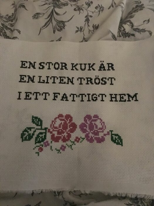 Broderi med texten "EN STOR KUK ÄR EN LITEN TRÖST I ETT FATTIGT HEM" och två broderade blommor i rosa och lila.