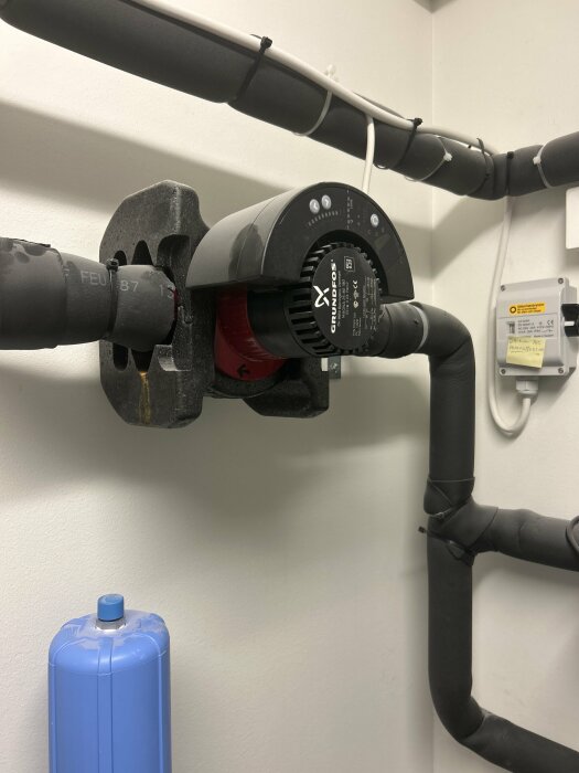 Vattenpump av märket Grundfos installerad i ett rörsystem med isolerade rör och en blå trycktank i bakgrunden.