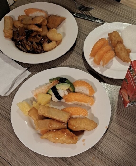 Tallrikar med buffémat inklusive sushi, vårrullar, friterad mat och en jordgubbsdryck på ett bord.