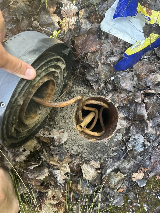 En hand pekar mot en öppning med rör som kan vara en pump, omgiven av löv och plastförpackningar.