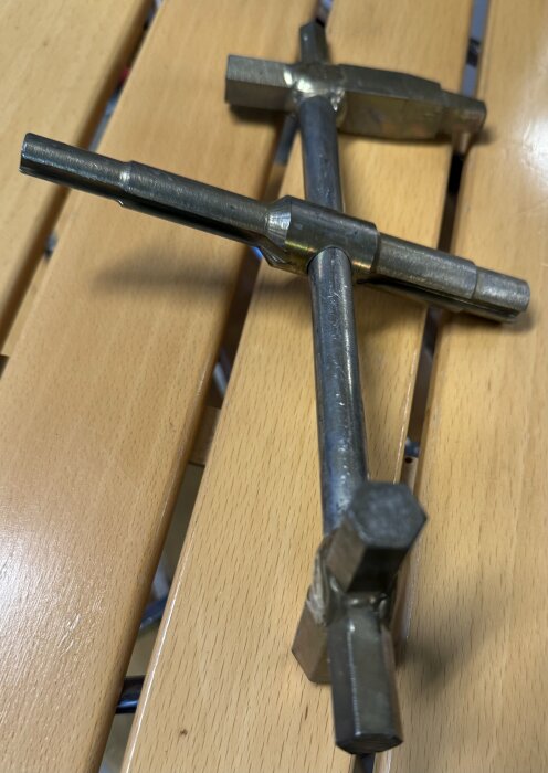Metallverktyg med korsformad konstruktion och rörliga delar på en stång, placerat på ett träbord. Funktionen är okänd för inläggsförfattaren.