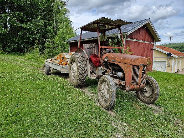 En gammal traktor med trästockar på en vagna i en grönskande trädgård, framför en röd lada och ett gult hus.