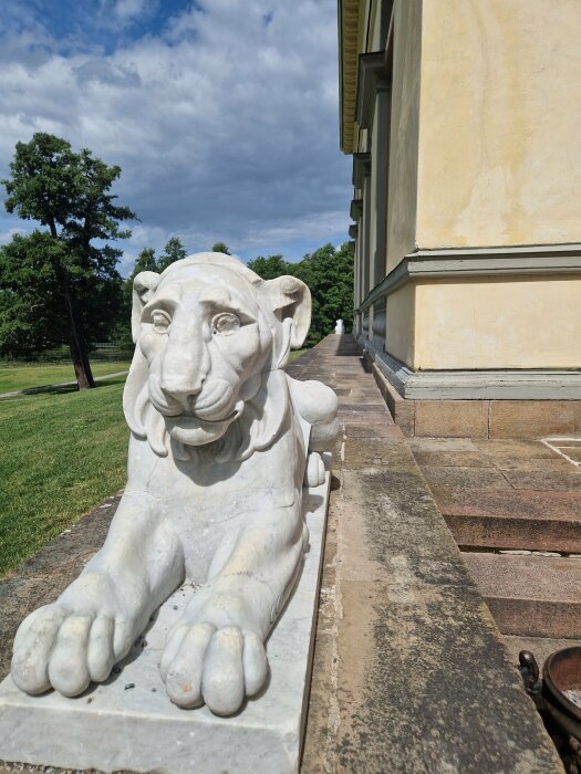 Staty av en liggande lejon vid en byggnads fasad, med gröna träd och blå himmel i bakgrunden.