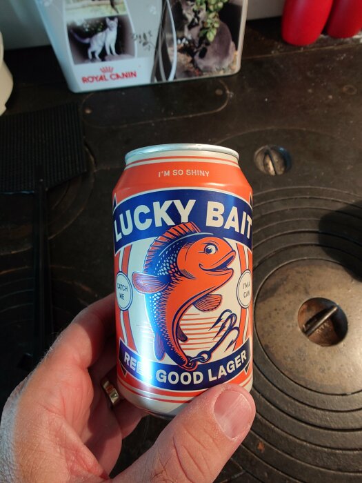 En hand håller en ölburk av märket Lucky Bait, med en bild på en fisk och texten "Reel good lager" på en köksbänk med en kattmatsburk i bakgrunden.