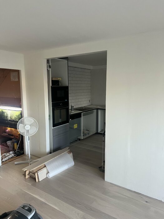 En öppning mellan ett kök med vita kakelväggar och vardagsrum, med några byggmaterial på golvet och en fläkt nära väggen.