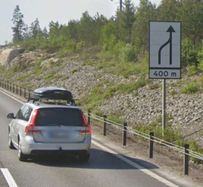 Vit kombibil med takbox kör på en landsväg med en skylt som visar att en uppfart kommer om 400 meter. Skog och berg i bakgrunden.