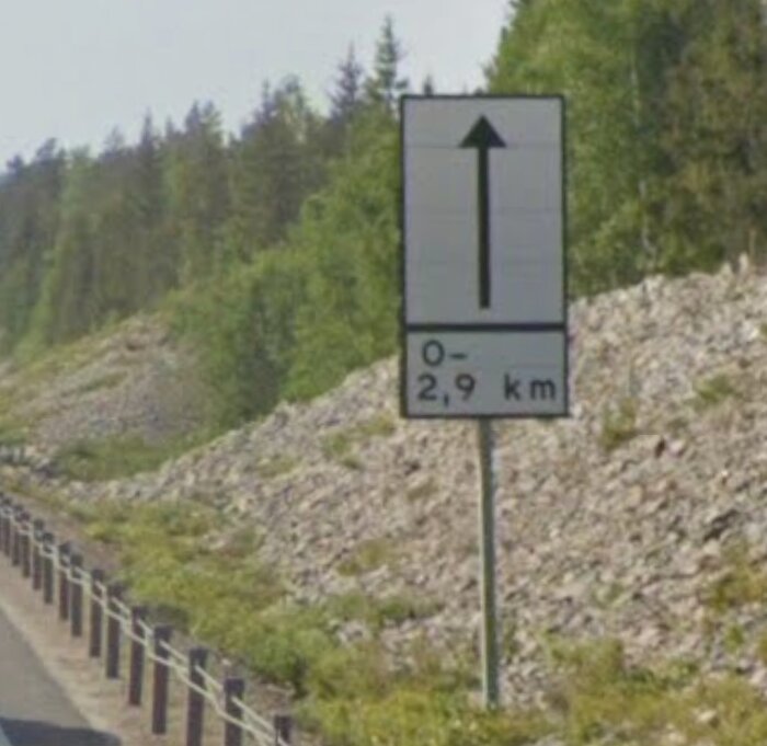 Vägmärke längs vägen som visar en pil uppåt med texten "0–2,9 km", omgiven av skog och stenar.