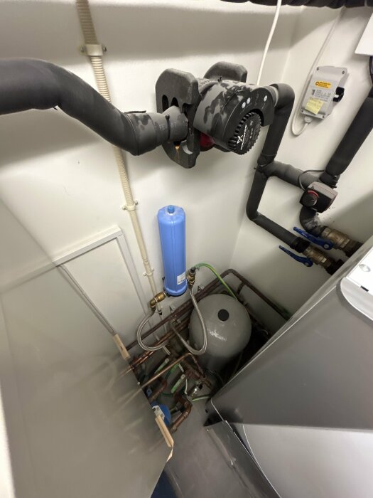 Notering av rör och komponenter i ett maskinrum för VVS-installation, inklusive en blå vattenfilter, svarta isolerade rör och en grå trycktank.
