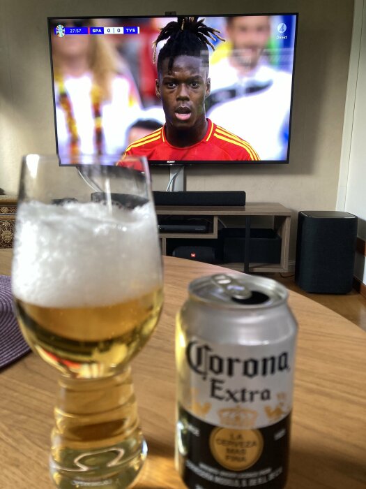 Ett glas öl och en ölburk står på ett bord framför en TV som visar en fotbollsmatch mellan Spanien och Tyskland.