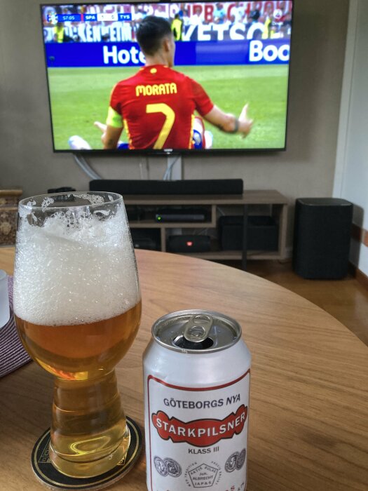 En ölburk och ett upphällt glas med Göteborgs Nya Starkpilsner på ett träbord framför en TV som visar en fotbollsmatch med en spelare i röd tröja och nummer 7.