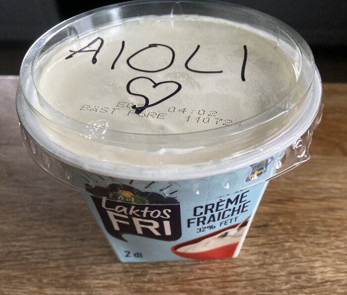 En burk laktosfri crème fraîche på ett träbord, med ett plastlock där ordet "AIOLI" är skrivet med en penna.